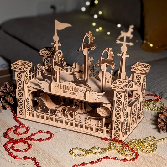 3D wooden “Reindeer Flight School” scene made from a laser cut digital file, shown assembled on a table with pinecones and holiday beads. Layered reindeer figures appear to practice flying on raised poles and platforms