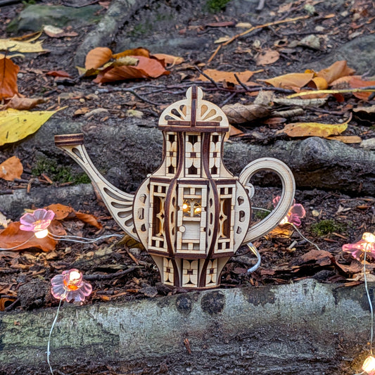 Laser cut wooden teapot fairy house displayed outdoors on tree roots, surrounded by fallen leaves and small glowing flower lights.