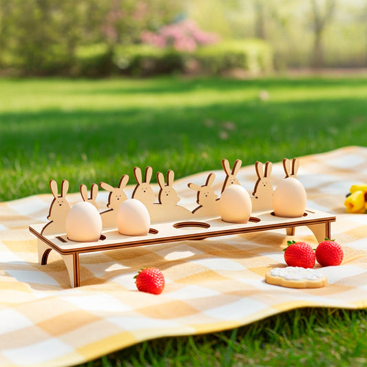 A natural wood laser plywood cut Easter egg holder shaped with bunny ear silhouettes, displaying light brown eggs on a yellow plaid picnic blanket with strawberries and a cookie