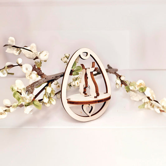 Laser cut plywood wooden Easter egg ornament featuring two kissing bunnies and a small heart on the base, framed by white blooming spring tree branches on a clean white background