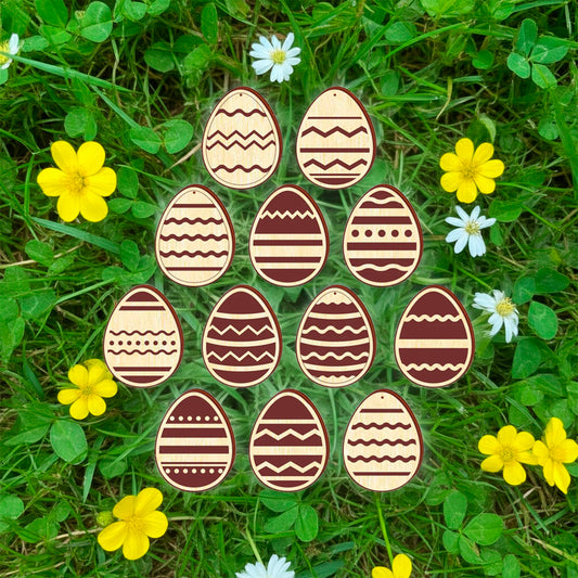 A set of twelve light brown laser cut wooden plywood Easter egg ornaments, each engraved with a different geometric or floral pattern, lying on a bright green grass background among yellow buttercups and small pink flowers