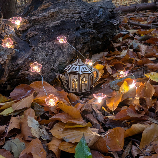 A miniature wooden fairy house shaped like a teapot, made from laser cut plywood, placed among autumn leaves with small glowing flower lights surrounding it.