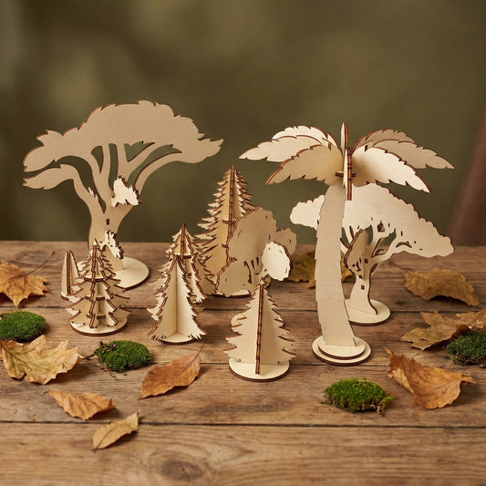 Collection of various wooden tree models crafted from laser cut plywood. The set features different heights and species, from spreading oaks to pointed firs. Displayed in a forest-like arrangement with real moss and dried leaves.