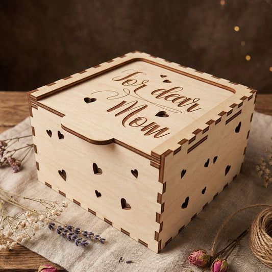 A close-up shot of a closed wooden gift box made from light-colored laser-cut plywood. The lid is engraved with "For dear Mom" and small heart accents. The sides of the box feature a pattern of tiny heart cutouts, highlighting the precision of the laser engraving. Posed on a linen cloth with dried lavender and rosebuds.
