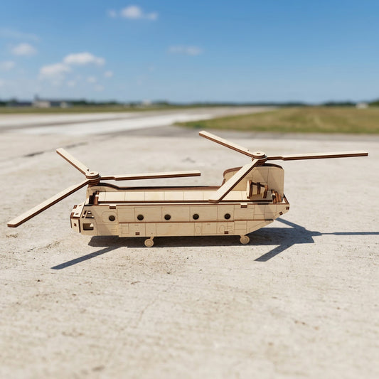 Side profile of a laser cut plywood cargo helicopter model. The long fuselage shows detailed etched panels, small landing wheels, and two sets of rotor blades. Displayed on a sunny airfield under a clear blue sky.