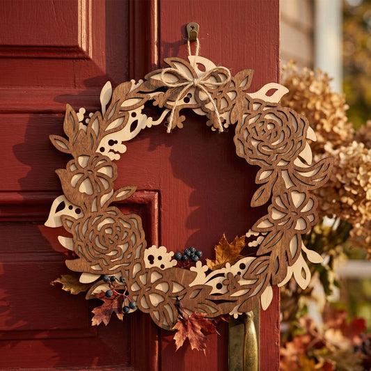 A handcrafted layered laser-cut plywood flower wreath hanging on a red wooden door. The wreath features intricate roses and foliage in contrasting light and medium brown wood tones. A simple twine bow is tied at the top, accented by real autumn leaves and berries for a rustic fall aesthetic.