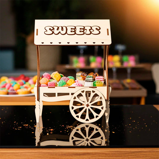 A light wooden plywood laser cut miniature vendor cart labeled "SWEETS" on the roof, filled with colorful jelly candies, resting on a reflective black surface with glitter, taken in bright outdoor lighting.