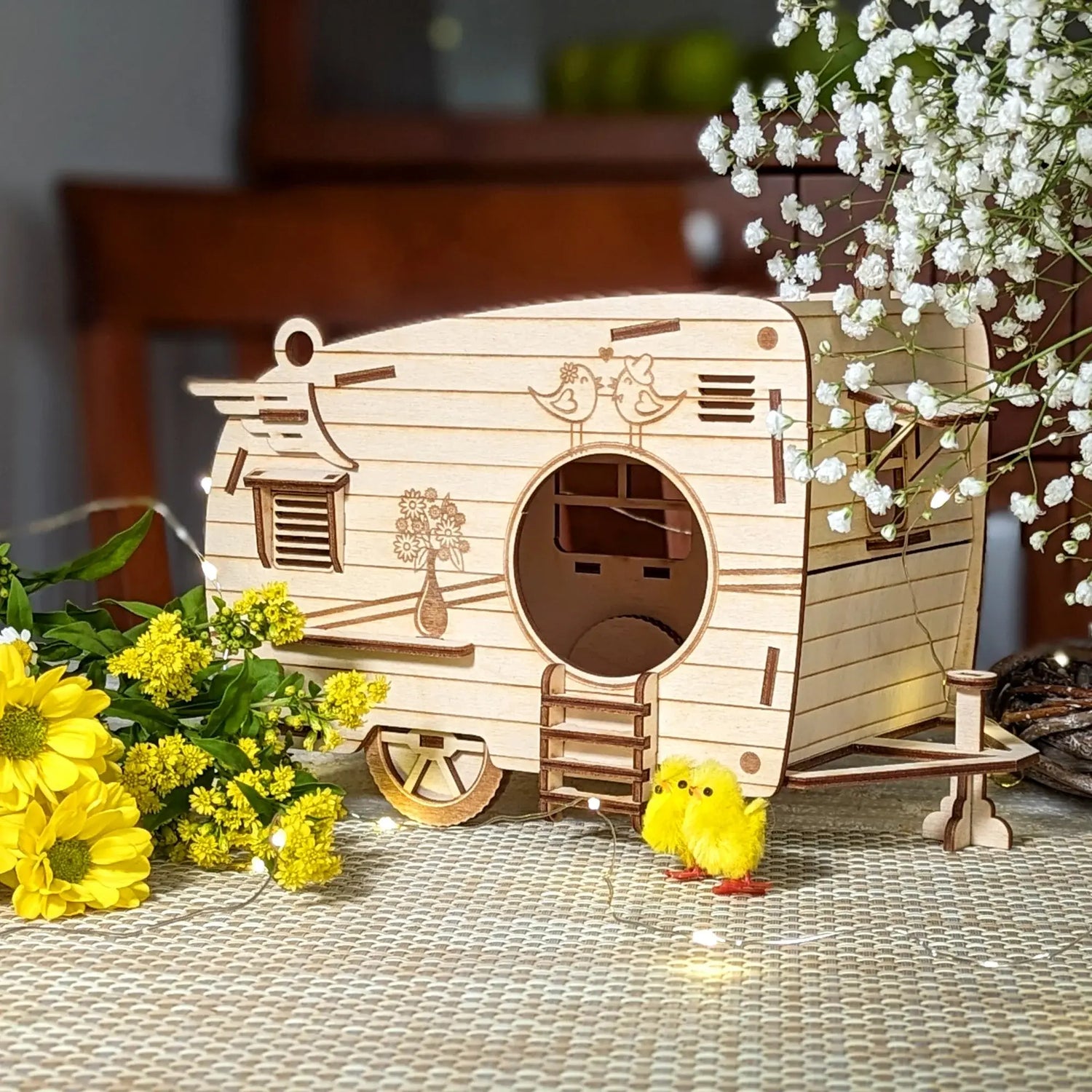 Miniature retro camper trailer model made of laser cut plywood, decorated with yellow flowers, tiny fuzzy chick figurines, and white baby's breath blossoms.