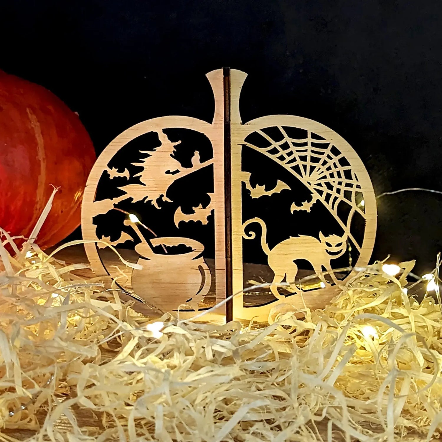 Laser cut plywood pumpkin ornament featuring spooky Halloween silhouettes of a witch flying over a cauldron, bats, a spiderweb, and a creepy cat, surrounded by straw and fairy lights.