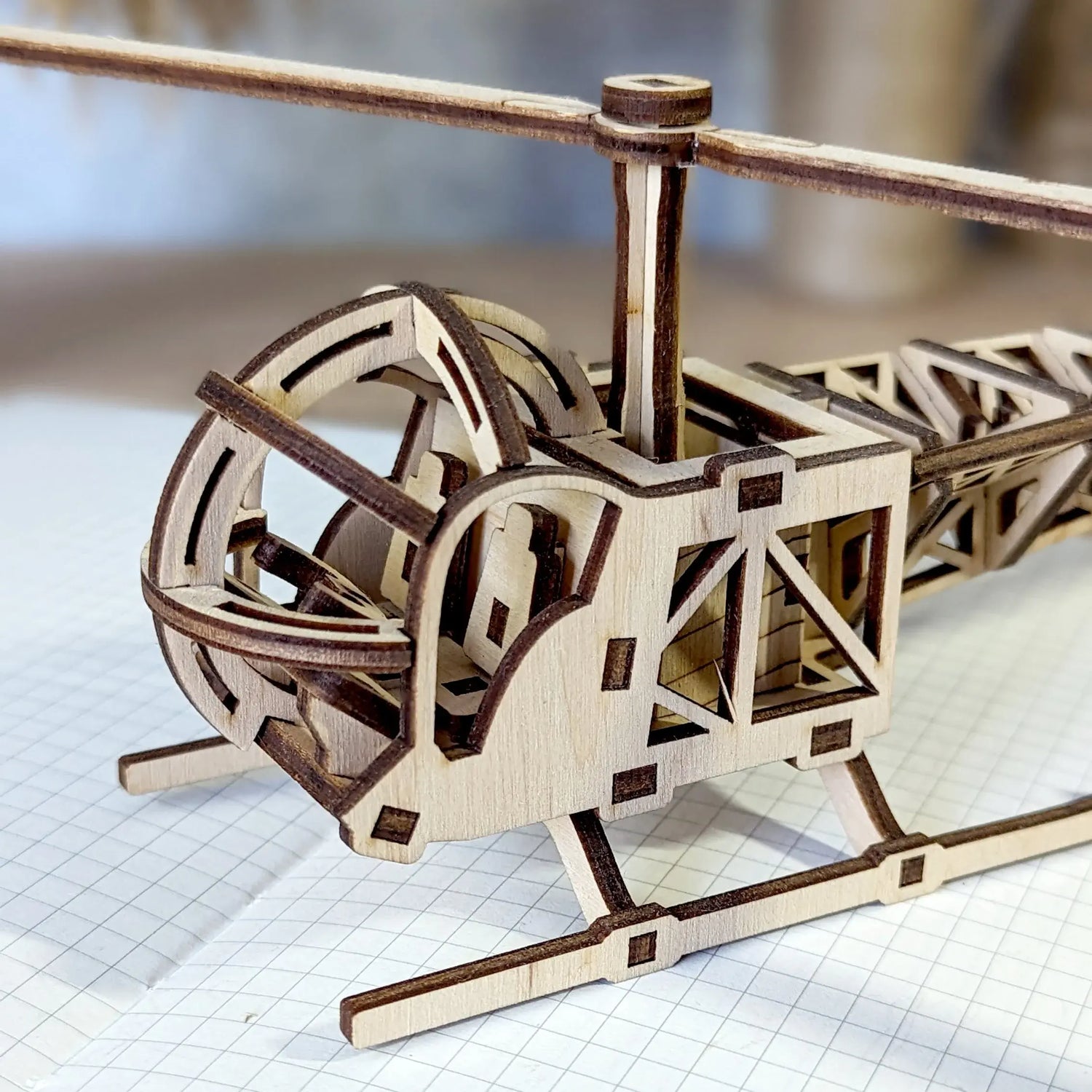 Miniature toy helicopter model made of laser cut plywood, showcasing detailed rotor blades and landing skids on graph paper.