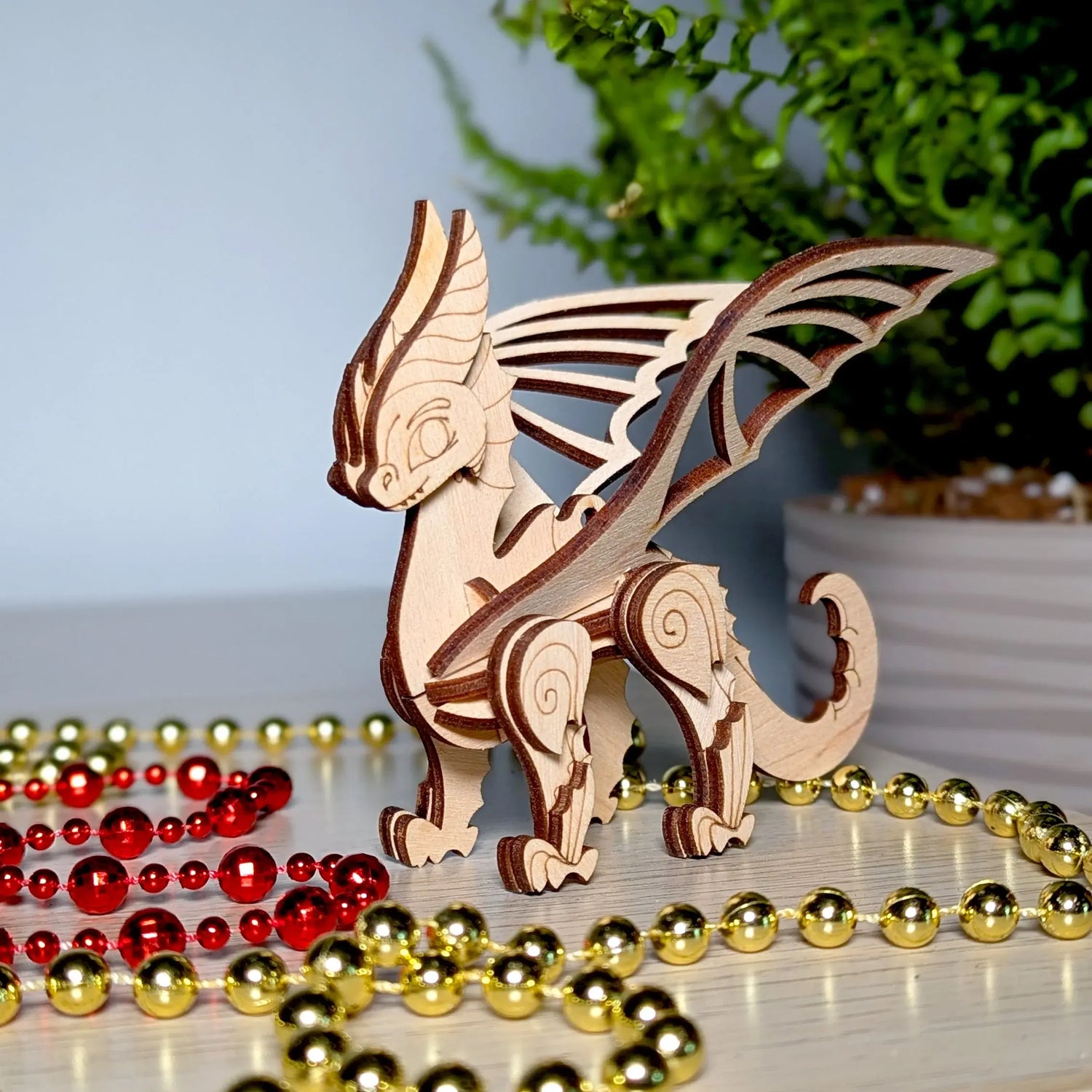 Miniature dragon figurine crafted from laser cut plywood, standing on a table with shiny red and gold bead garlands.