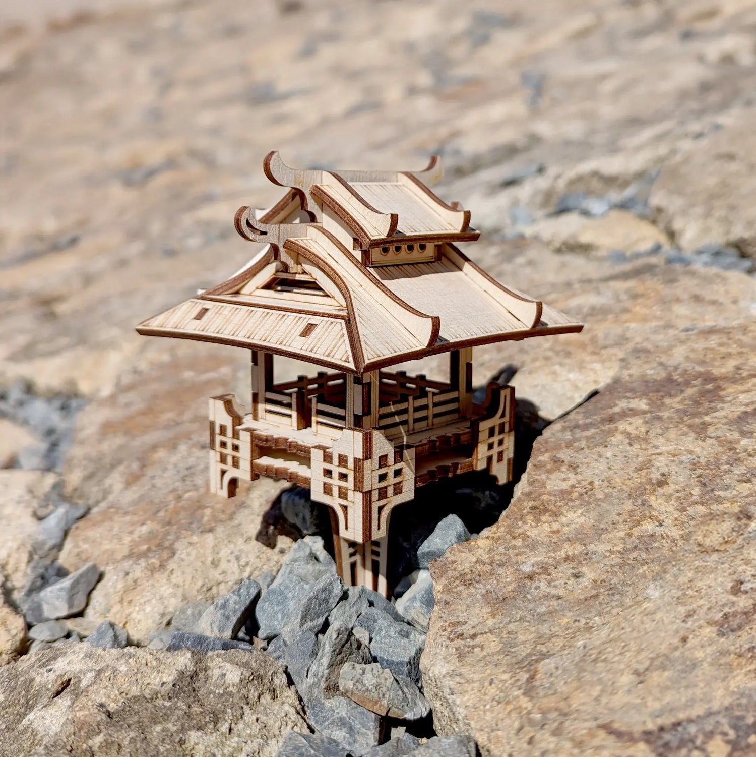 Miniature Asian pagoda pavilion crafted from laser cut plywood, placed outdoors on rugged natural stones and gravel.