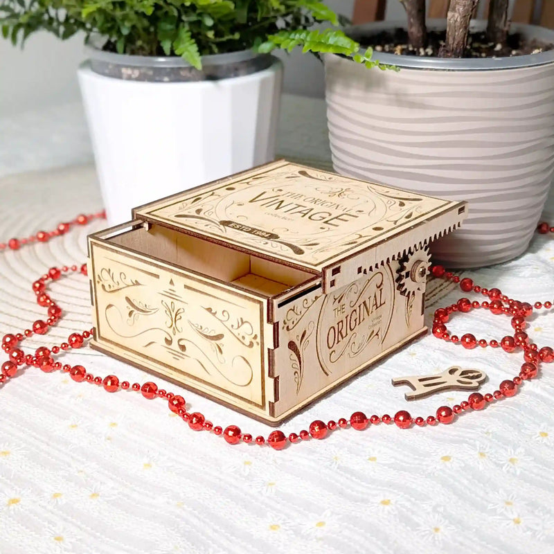 Vintage-style laser-cut wooden jewelry box with an open drawer and decorative engravings, placed on a table with red beads and a small wooden key, with potted plants in the background.