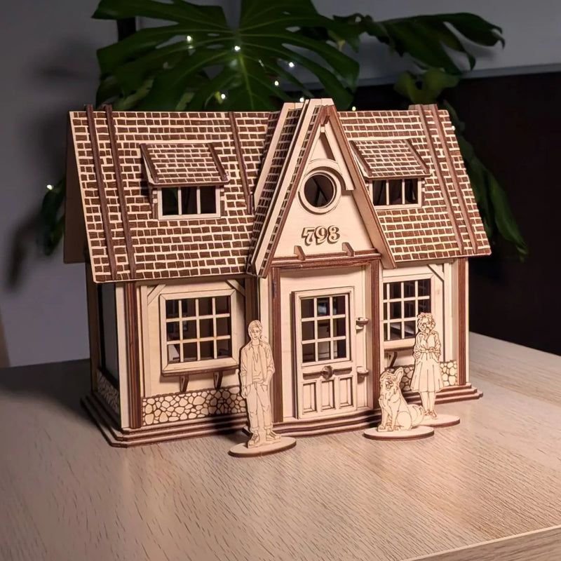 Dollhouse laser-cut wooden model of a cozy house with engraved brick texture and roof tiles, displayed on a table with small wooden figures of people and a dog in front, and soft lights and a plant in the background.