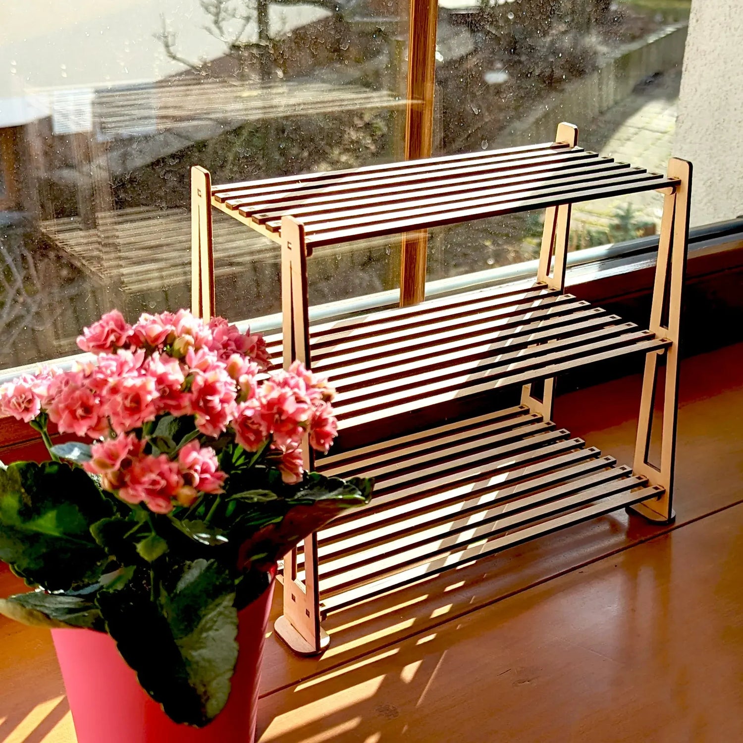 Empty three-tier slatted shelf made of laser cut plywood, placed on a sunlit windowsill next to a pink potted flower.