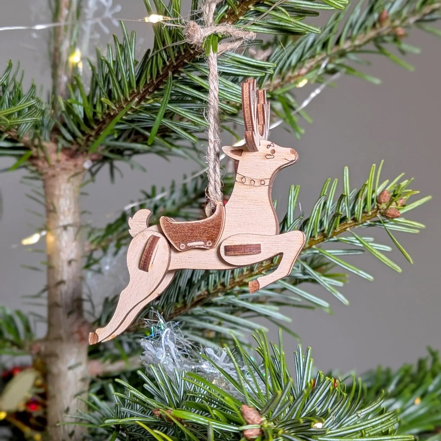 Running deer ornament made of laser cut plywood with twine string, hanging as a decoration on a green pine tree branch.