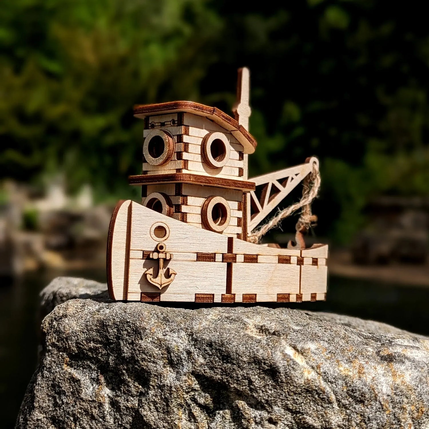 Miniature toy tugboat puzzle made of laser cut plywood, sitting outdoors on a large grey stone with a blurry forest background.