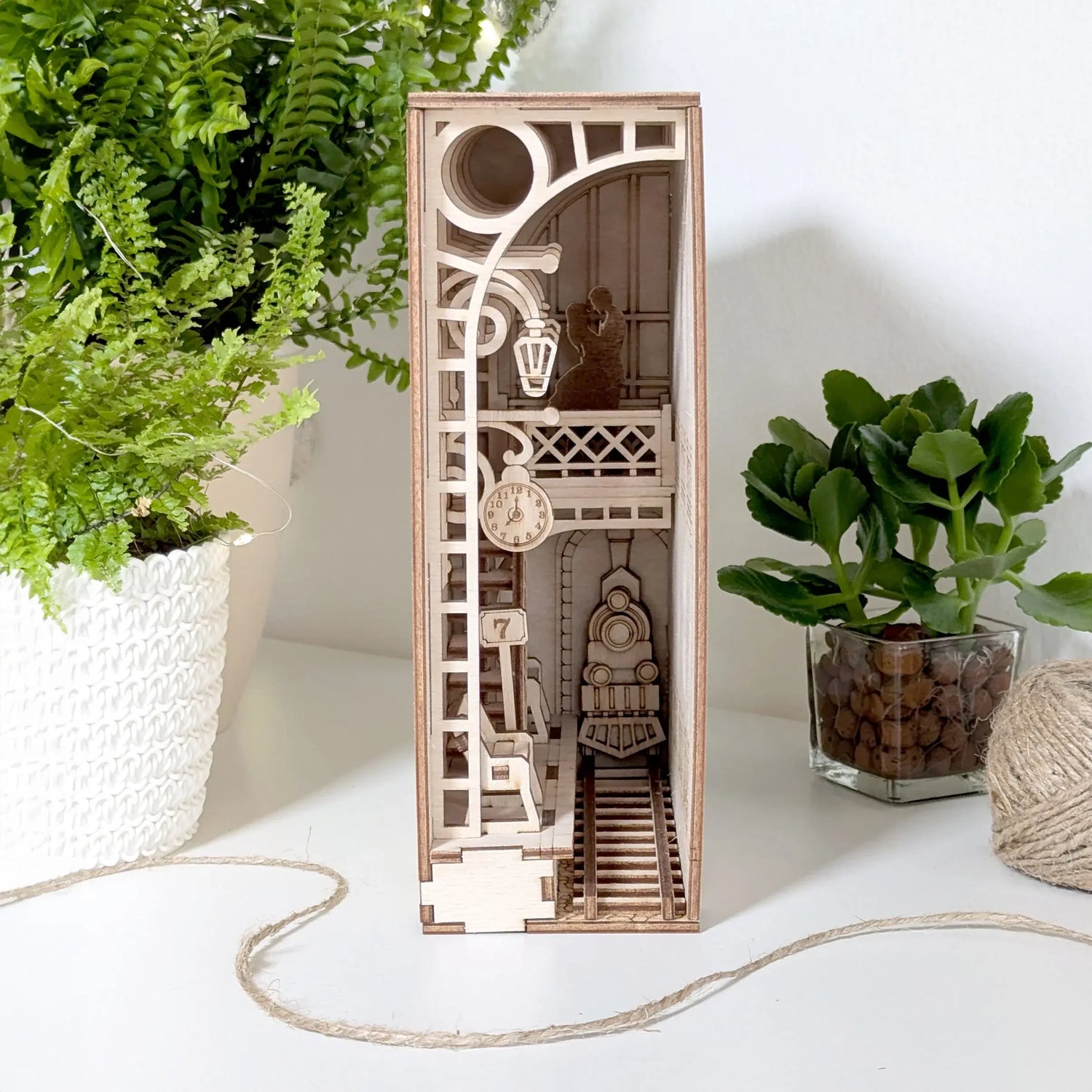 Vintage railway station book nook made of laser cut plywood, showing a tiny steam train on tracks beside home potted plants.