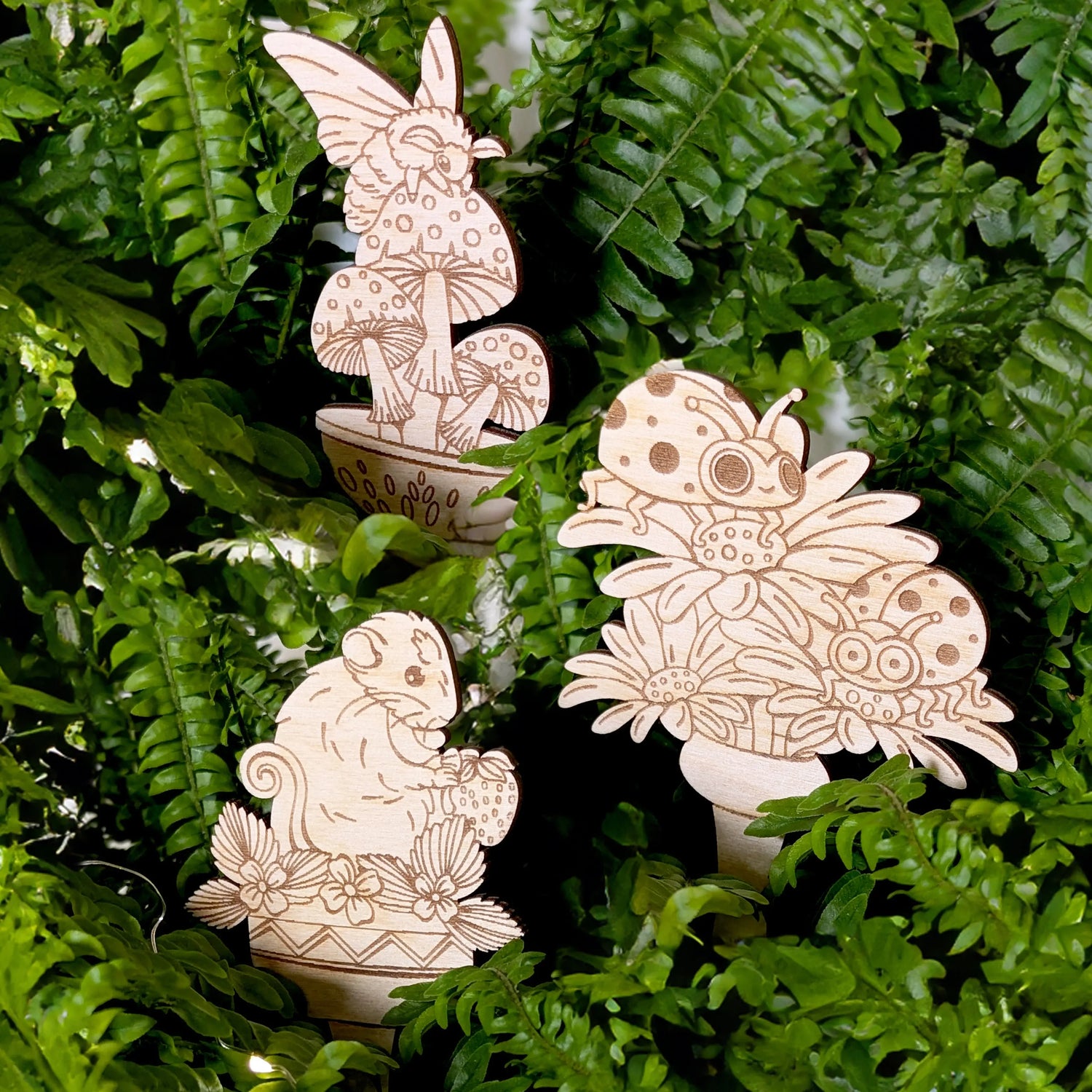 Whimsical garden stakes made of laser cut plywood with cute insect and animal carvings, tucked inside lush green fern leaves.