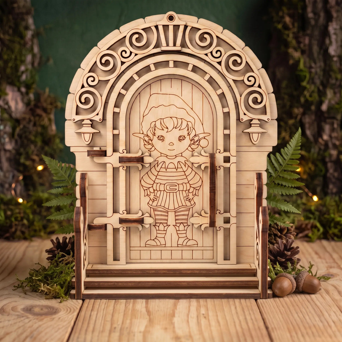 Front view of laser cut plywood elf door figurine with stairs, pinecones, and acorns on wooden table against green wall.