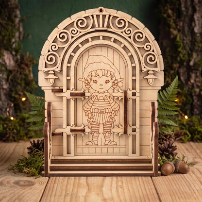 Front view of laser cut plywood elf door figurine with stairs, pinecones, and acorns on wooden table against green wall.