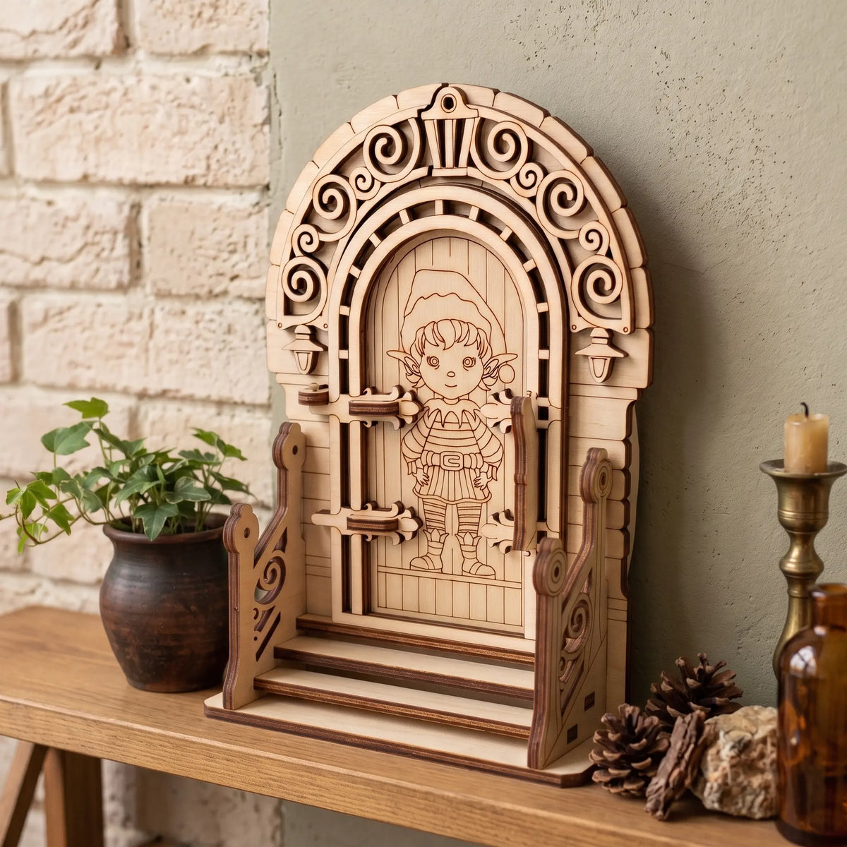Laser cut plywood fairy elf door with railings and steps standing on shelf next to potted ivy, candle, and pine cones.