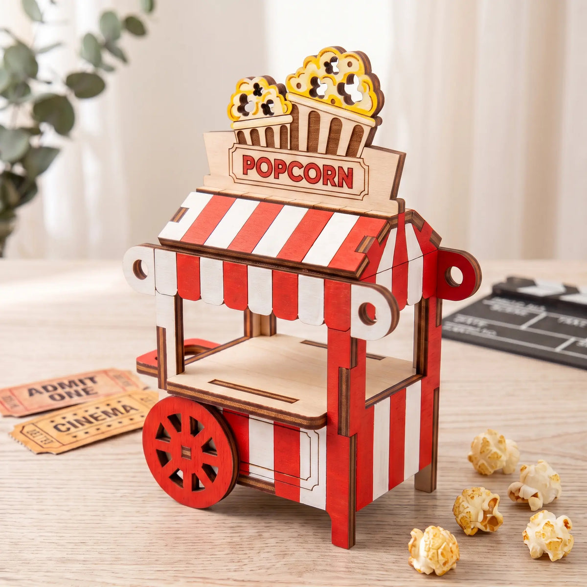 Painted red and white laser cut plywood popcorn cart model on wooden table with cinema tickets and popped popcorn kernels.