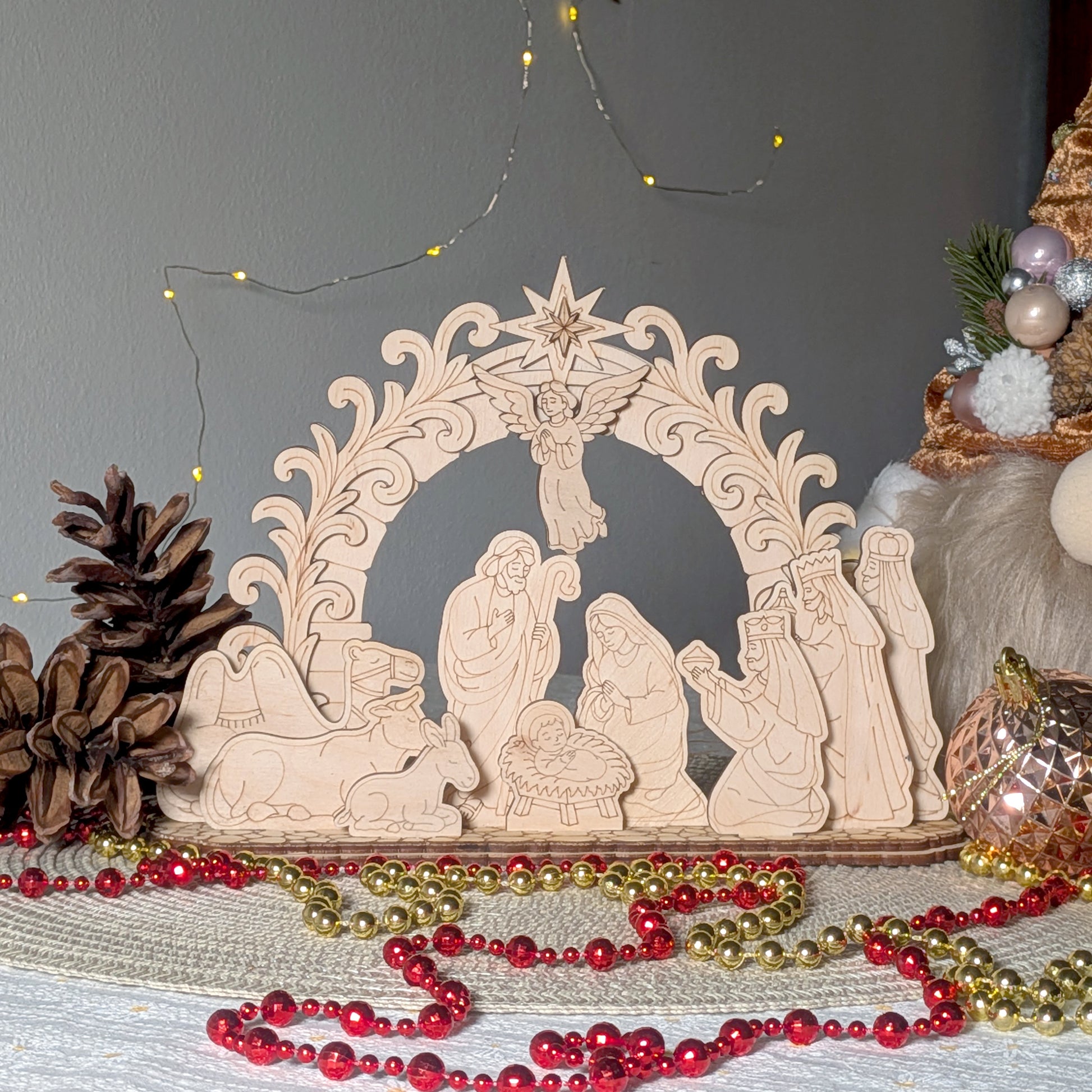 Laser cut wooden nativity scene displayed on a table, featuring Mary, Joseph, baby Jesus, shepherds, animals, and an angel under a decorative arch. Light wood texture with fine engraved details