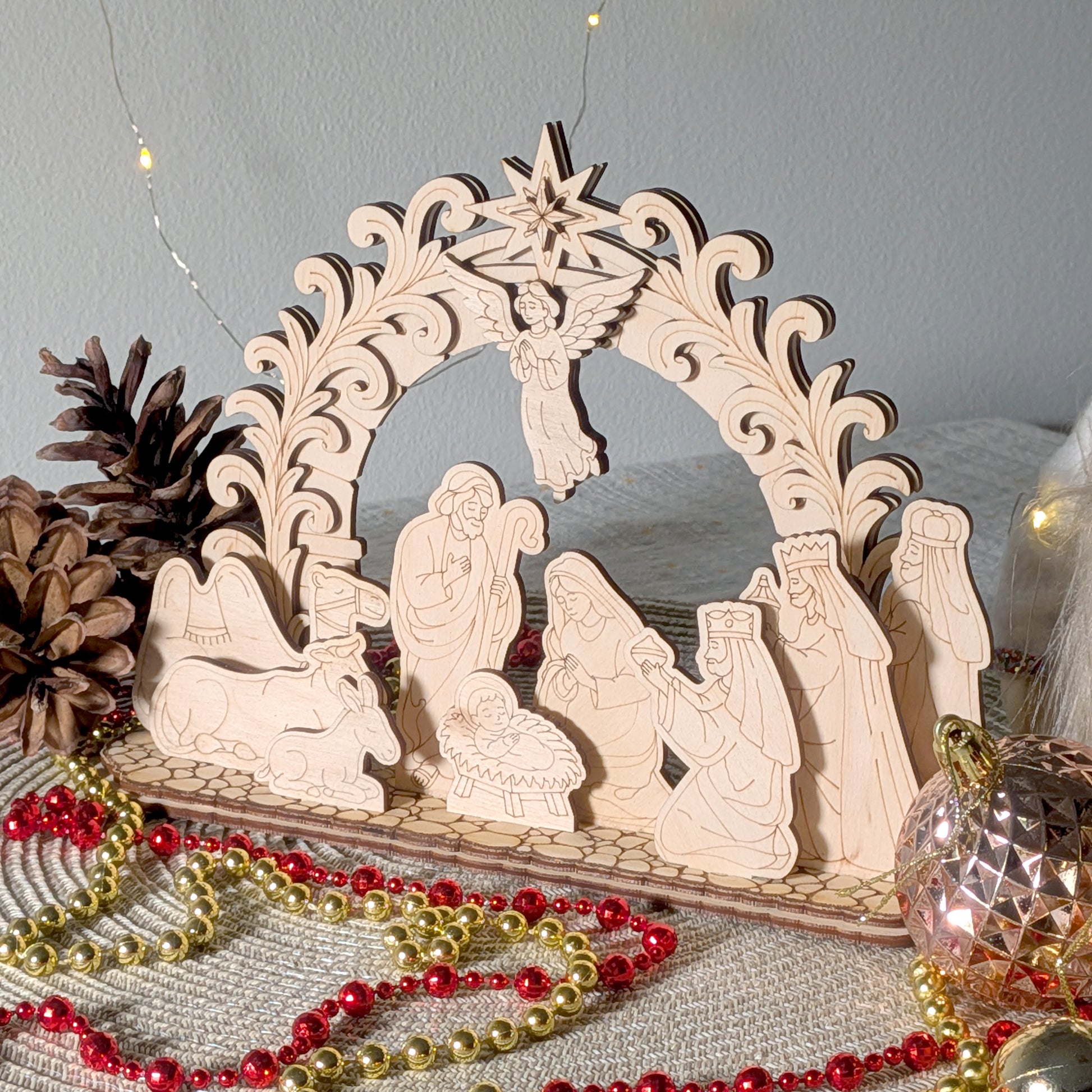 Laser cut wooden nativity scene set on a table, showing Mary, Joseph, baby Jesus, an angel, and the three wise men under a decorative arch. Light wood with fine engraved details, surrounded by pinecones and festive beads