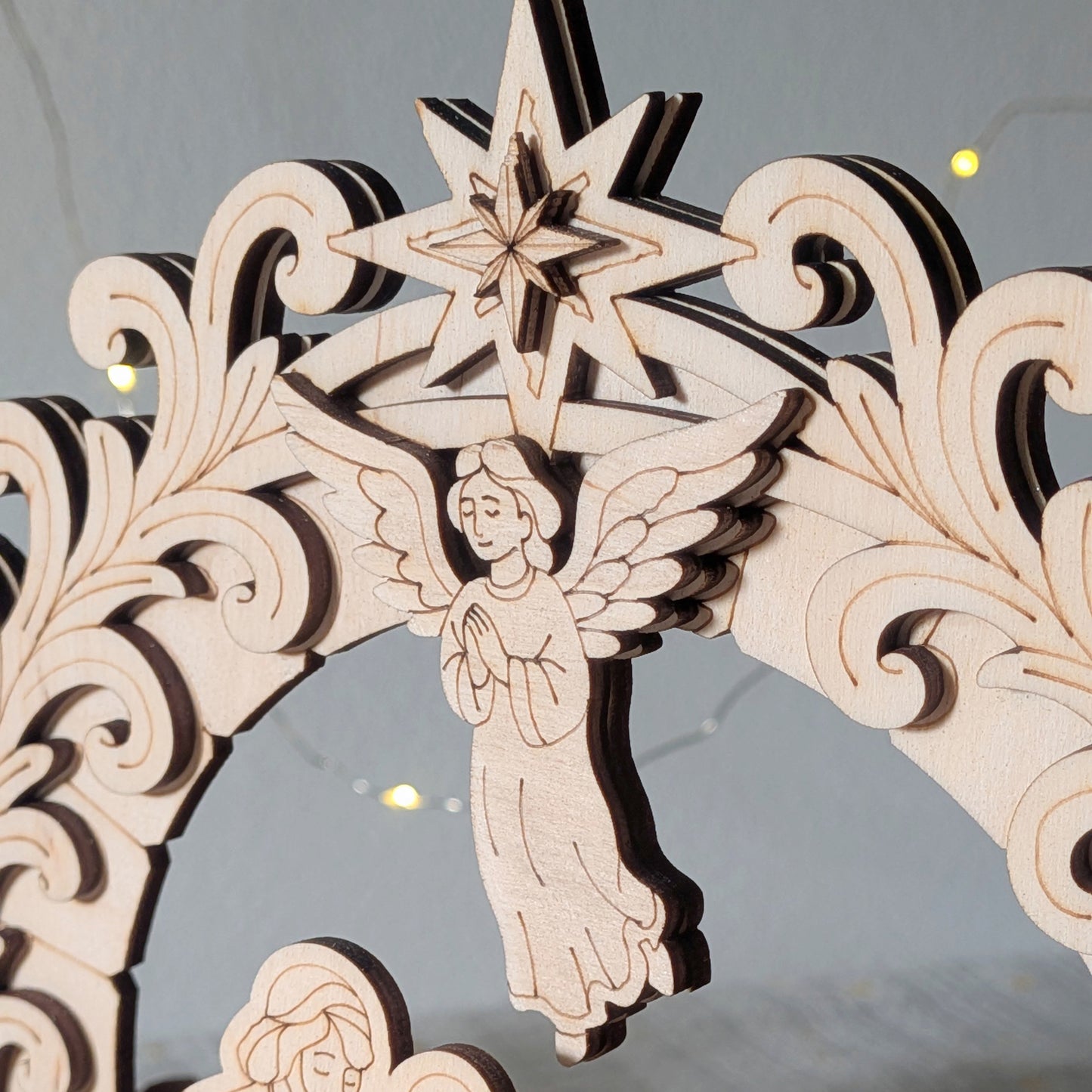 Close-up of a laser cut wooden nativity arch showing an angel with folded hands beneath a layered star. Light wood with engraved details and smooth curved decorative patterns