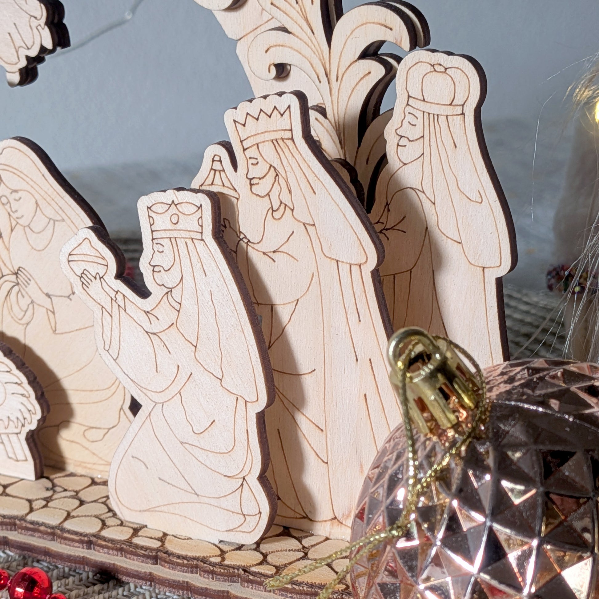 Close-up of laser cut wooden figures of the three wise men presenting gifts, with detailed engraved robes and crowns. Light wood texture, part of a nativity scene display, with a shiny ornament in the foreground