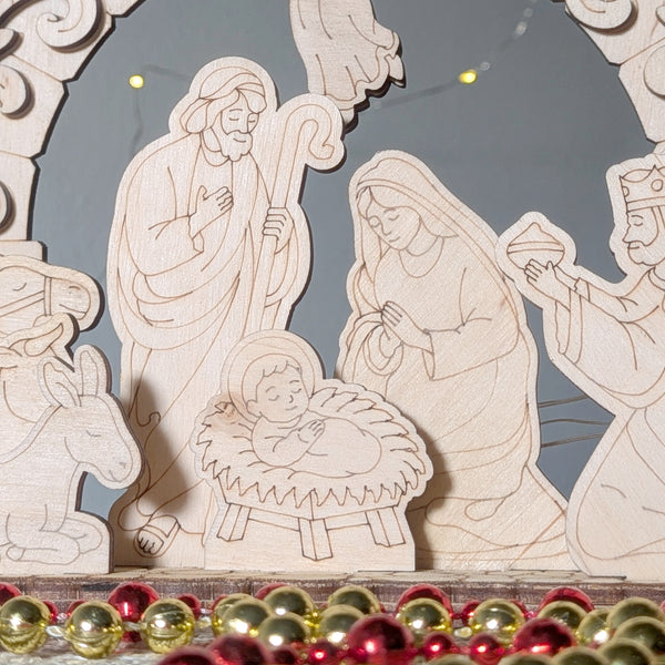 Close-up of a laser cut wooden nativity scene showing Mary, Joseph, and baby Jesus in a manger, with gentle engraved details and light wood texture. Decorative beads lie in front of the display