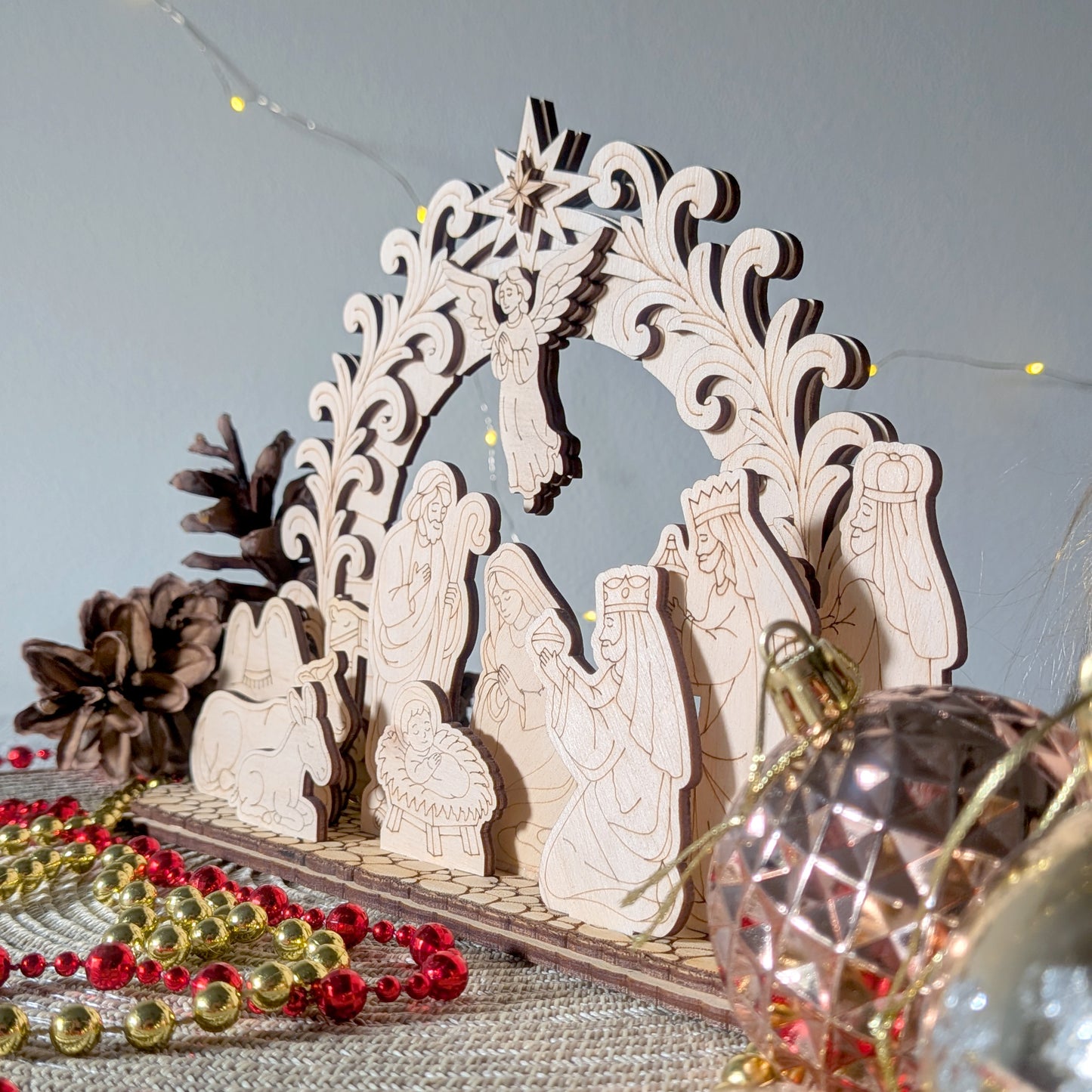 Side view of a laser cut wooden nativity scene with Mary, Joseph, baby Jesus, an angel, animals, and the three wise men under an ornate arch. Displayed with festive beads and pinecones
