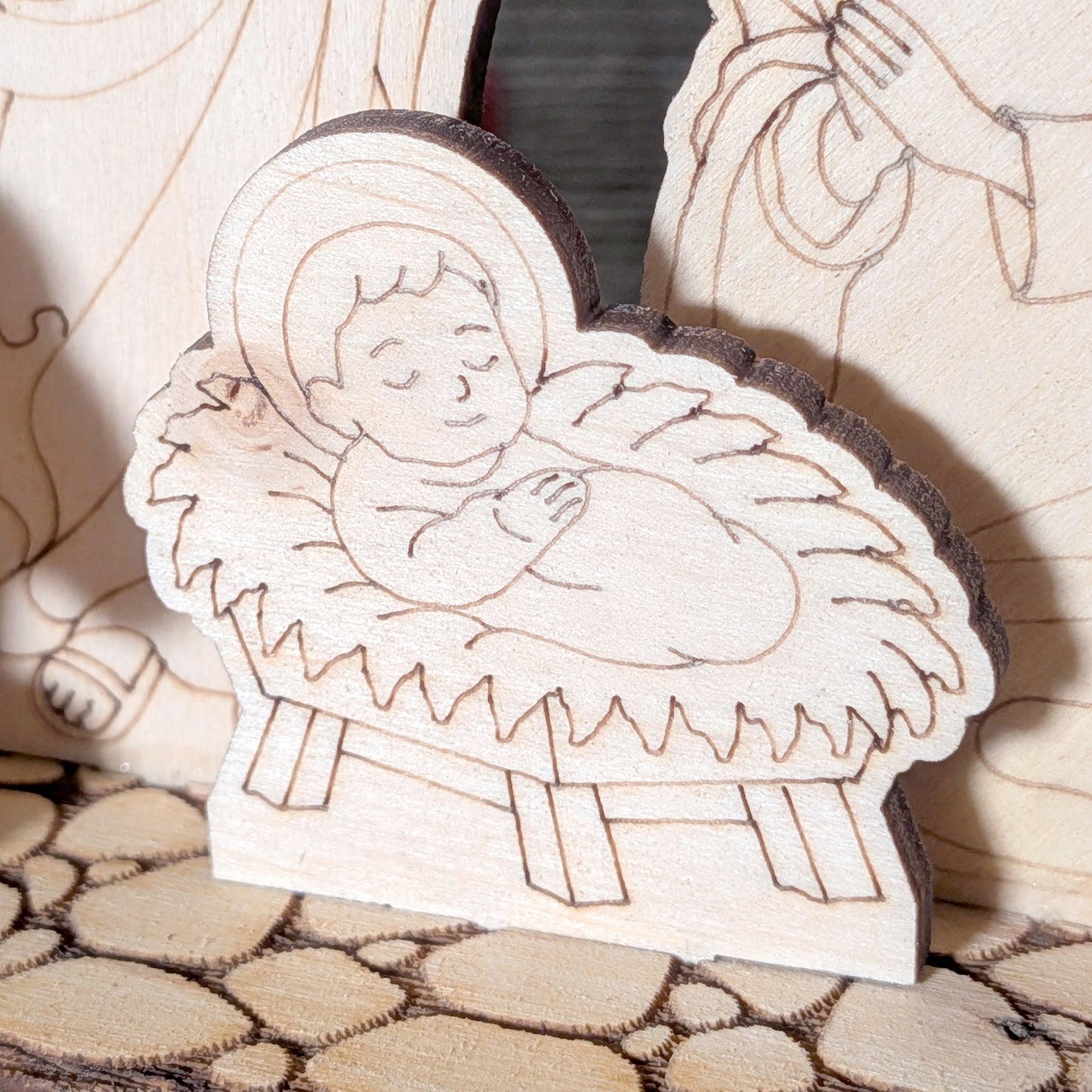 Close-up of a laser cut wooden baby Jesus figure lying in a manger, with engraved facial features and soft layered straw details. Light wood texture with smooth lines