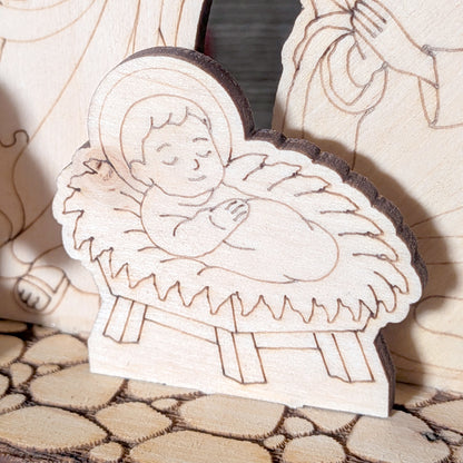 Close-up of a laser cut wooden baby Jesus figure lying in a manger, with engraved facial features and soft layered straw details. Light wood texture with smooth lines