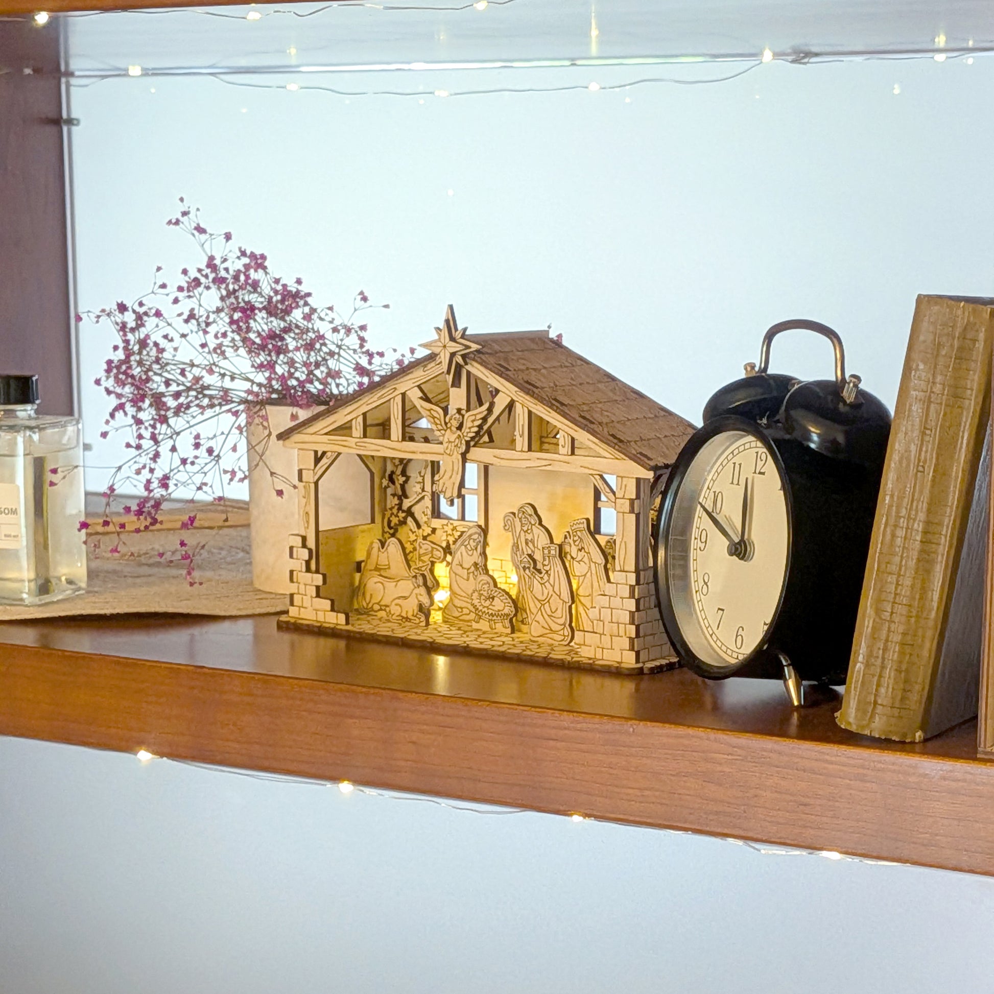 Wooden Nativity Scene with warm backlighting displayed on a shelf beside books, flowers, and a vintage clock. DIY Christmas decor created using a laser cut digital template