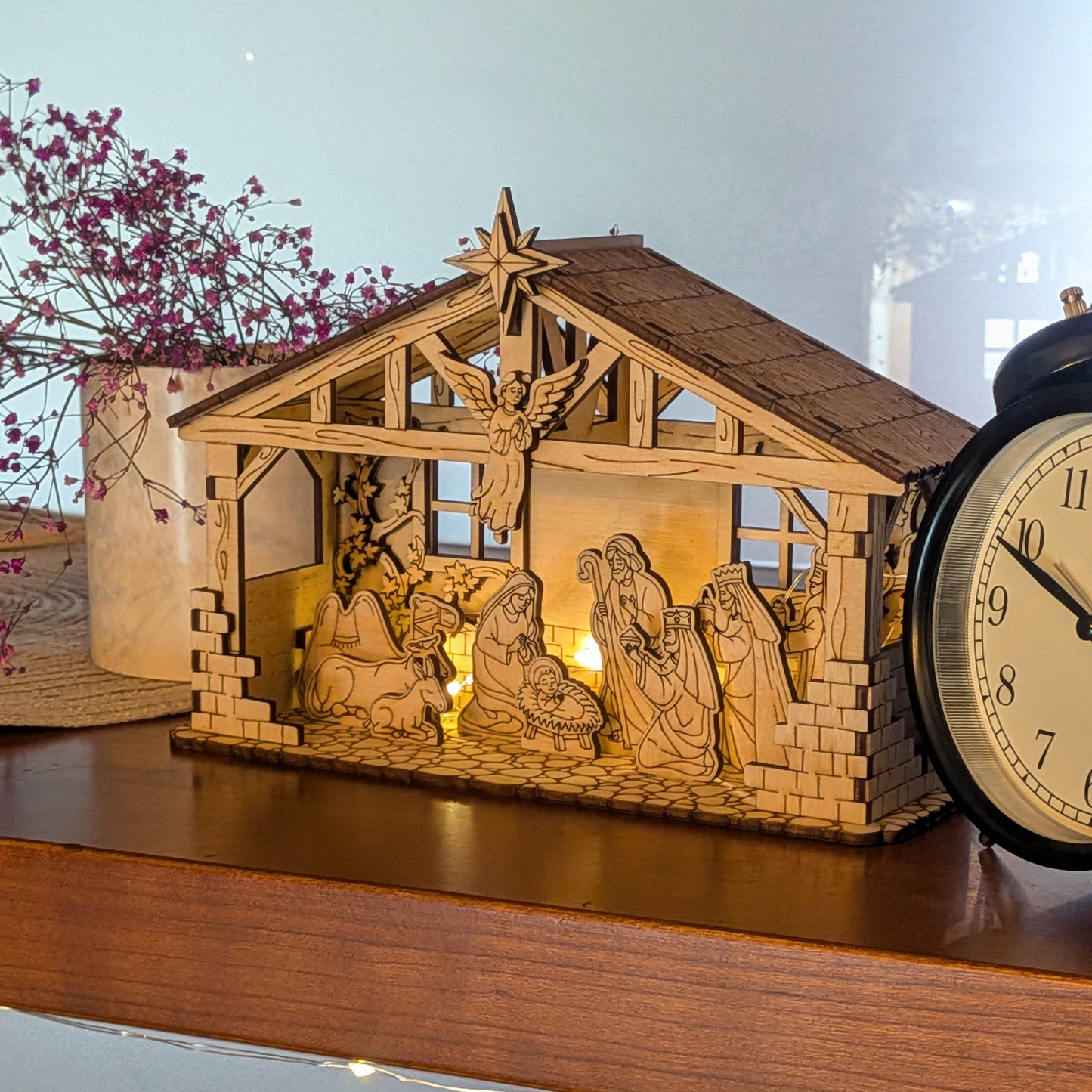 Wooden Nativity Scene displayed on a shelf with a clock and decorative flowers, featuring the Holy Family, angel, and Wise Men. Christmas decor made from a laser cut digital file template