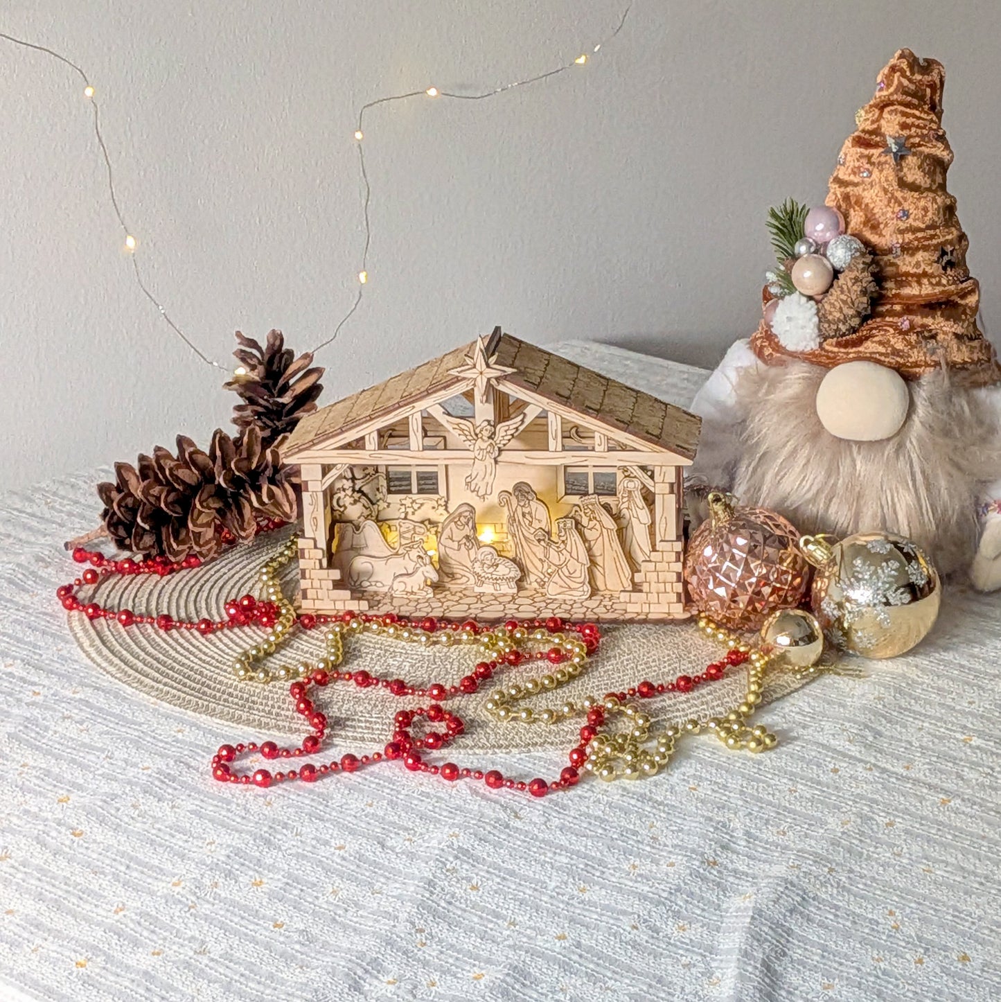 Wooden laser cut Nativity Scene with warm LED lights displayed on a holiday table with Christmas ornaments, pinecones, a festive gnome and red and gold bead garlands
