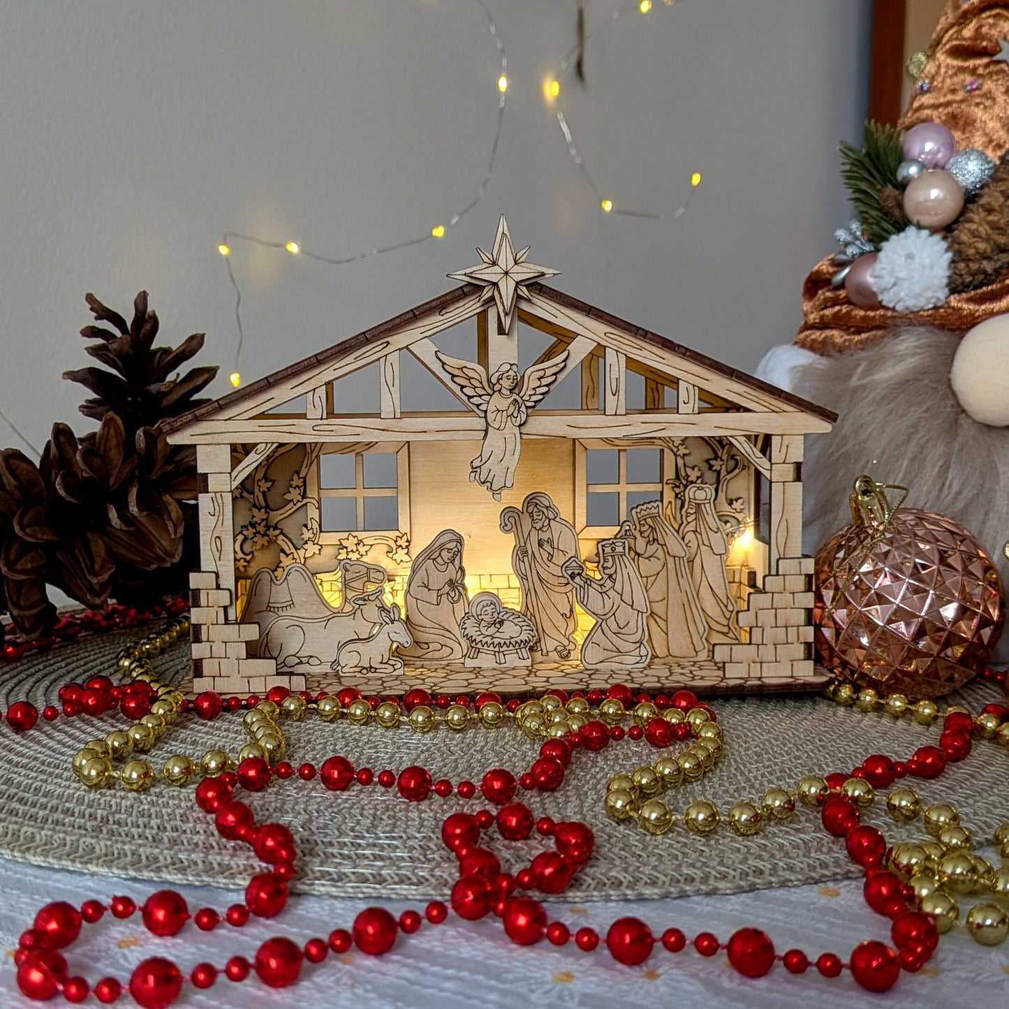 Wooden layered Nativity Scene with baby Jesus, Mary, Joseph, angel and the Three Wise Men inside a stable, displayed with Christmas ornaments and lights. Designed as a laser cut digital template for plywood