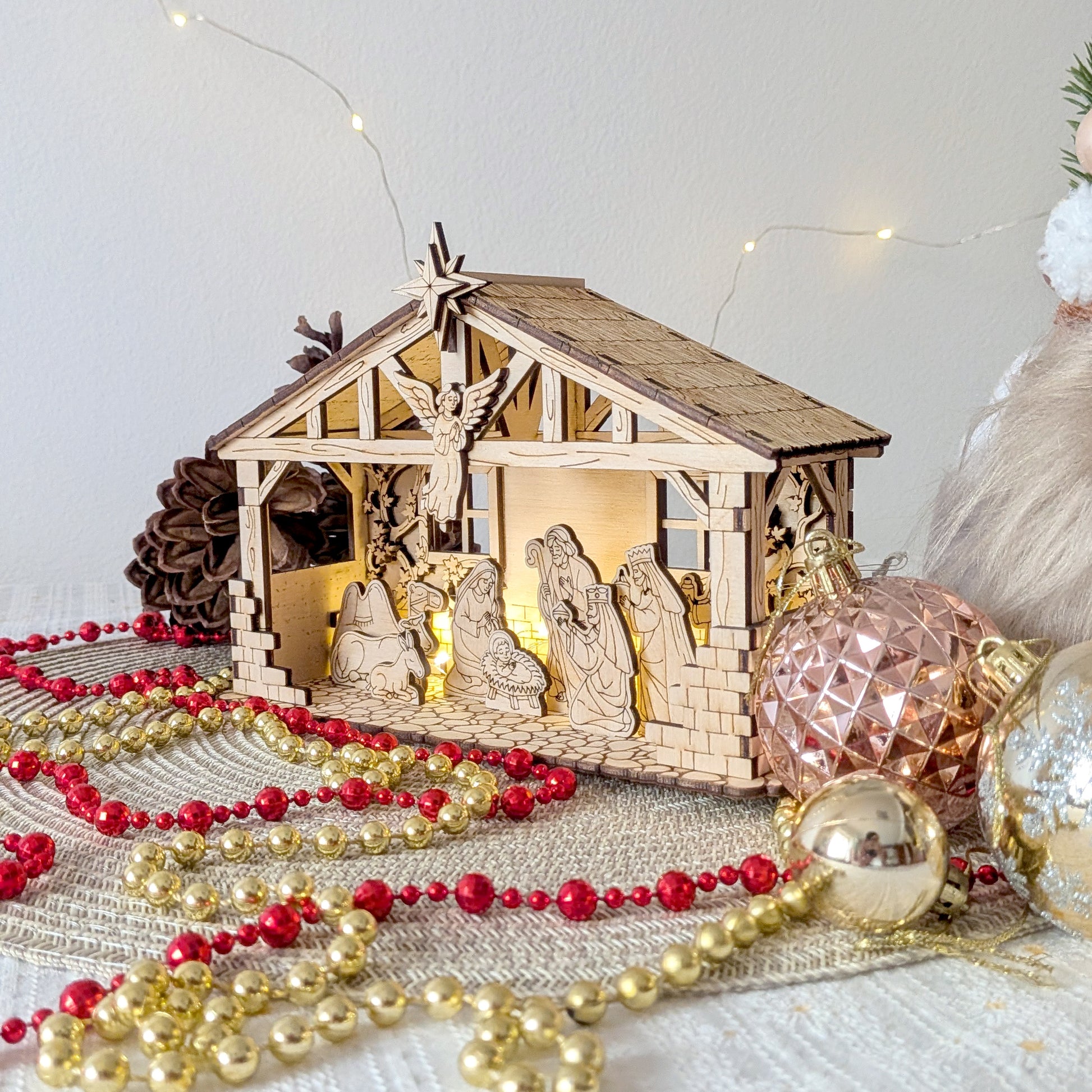 Wooden laser cut Nativity Scene with LED lights displayed on a festive Christmas table with ornaments and bead garlands. DIY holiday manger decoration featuring Mary, Joseph, Baby Jesus, angel and animals