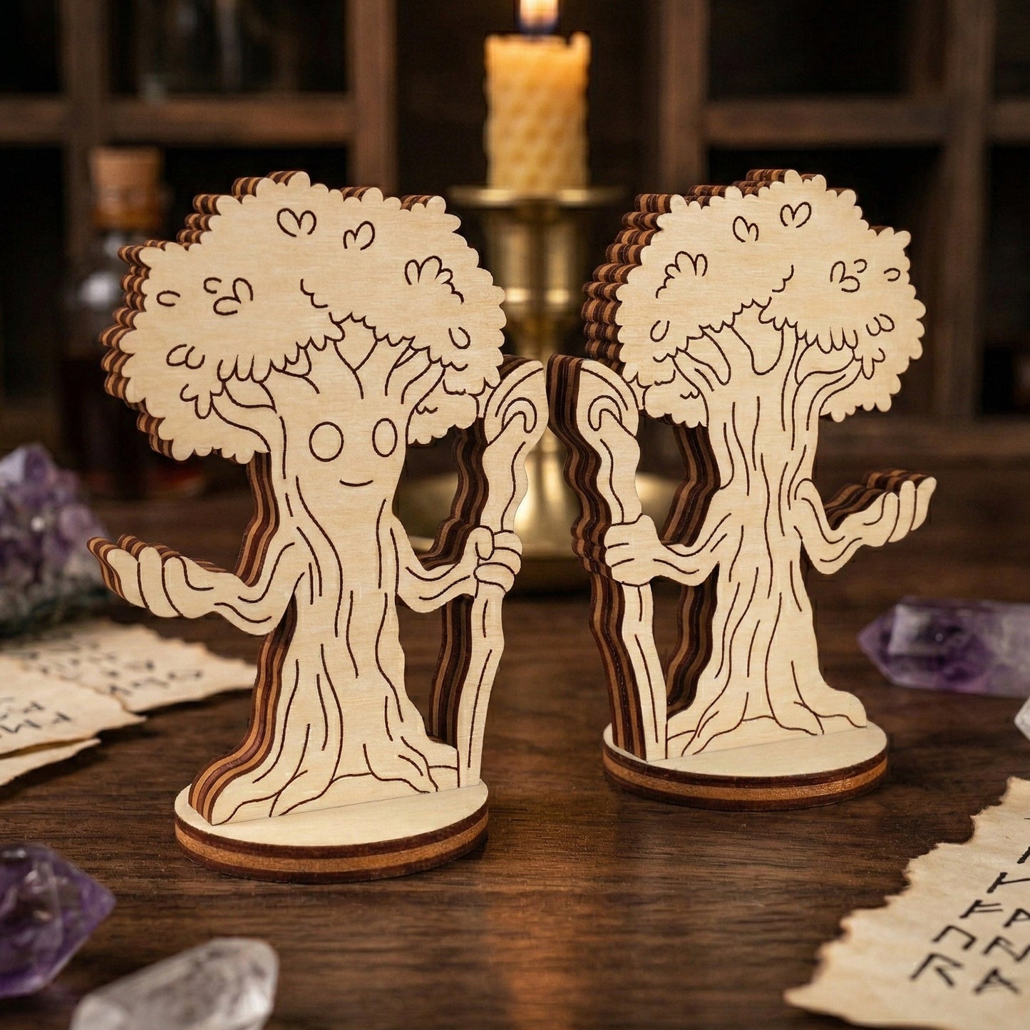 Two laser-cut plywood miniatures of a forest mage character, designed as a sentient tree with a friendly face and a staff. The wooden figurines are displayed on a dark surface alongside magical props like a lit candle, purple crystals, and runic papers for a mystical aesthetic.