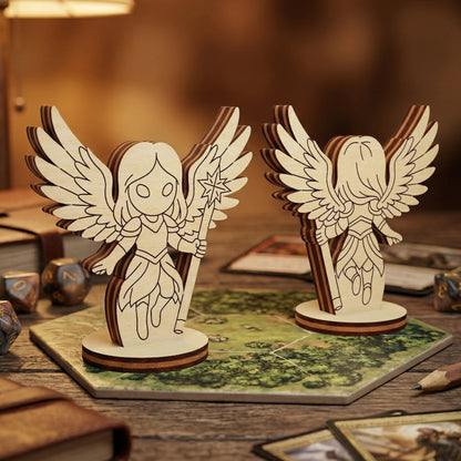 Front and back views of a 3D laser-cut plywood celestial angel figurine. The chibi-style angel character features intricately layered wings and a star-tipped staff. Displayed on a hexagonal tabletop gaming map surrounded by RPG accessories like polyhedral dice and cards in a cozy, fantasy-themed setting.
