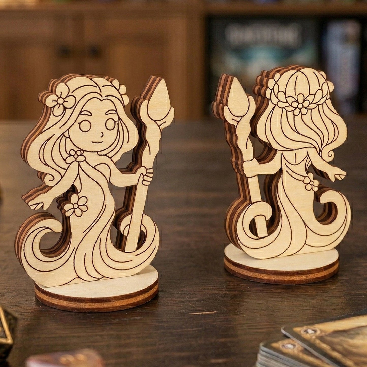 Front and back views of a 3D laser-cut plywood forest dryad figurine. The chibi-style character features flowing hair with flowers and holds a wooden staff. Displayed on a dark tabletop alongside polyhedral gaming dice and a stack of RPG cards in a cozy gaming environment.