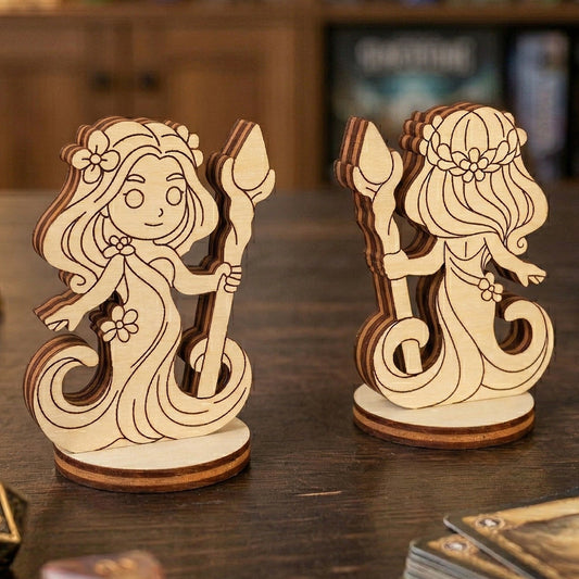 Front and back views of a 3D laser-cut plywood forest dryad figurine. The chibi-style character features flowing hair with flowers and holds a wooden staff. Displayed on a dark tabletop alongside polyhedral gaming dice and a stack of RPG cards in a cozy gaming environment.