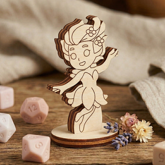 Close-up of a laser cut plywood fairy figurine with engraved details, standing on a round base among RPG dice and flowers.