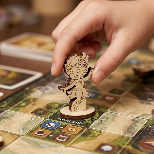 Hand placing a laser cut plywood fairy figurine on a "Forest path" board game tile, showing its use as a tabletop miniature.