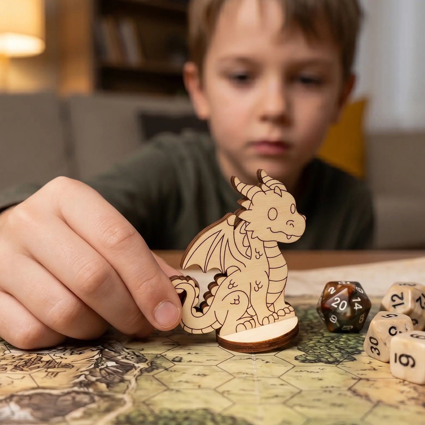 A child’s hand holds a laser cut plywood dragon miniature on a hex grid game map. Scale is shown next to standard gaming dice.