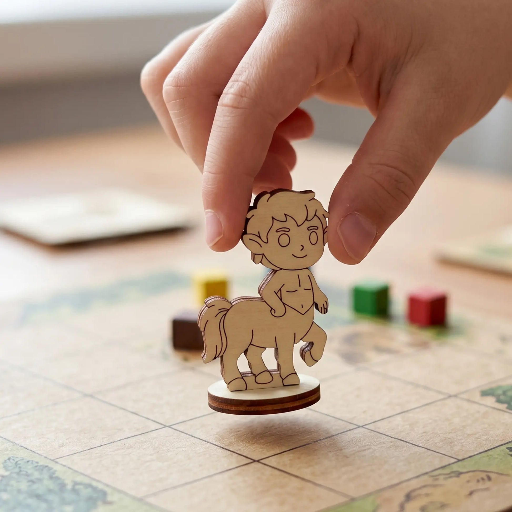 Close-up of a hand holding a layered laser cut plywood centaur figurine over a grid game board with small colorful wood cubes.