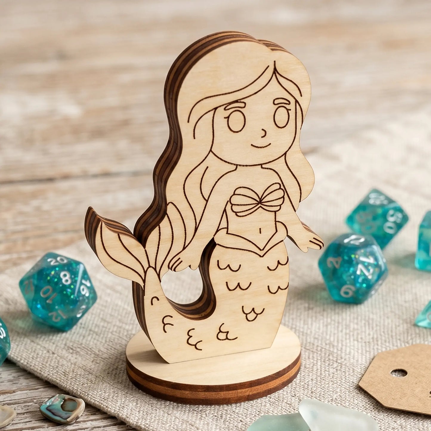 Detailed close-up of a laser cut plywood mermaid figurine with wood grain layers, standing on linen near teal glittery dice.