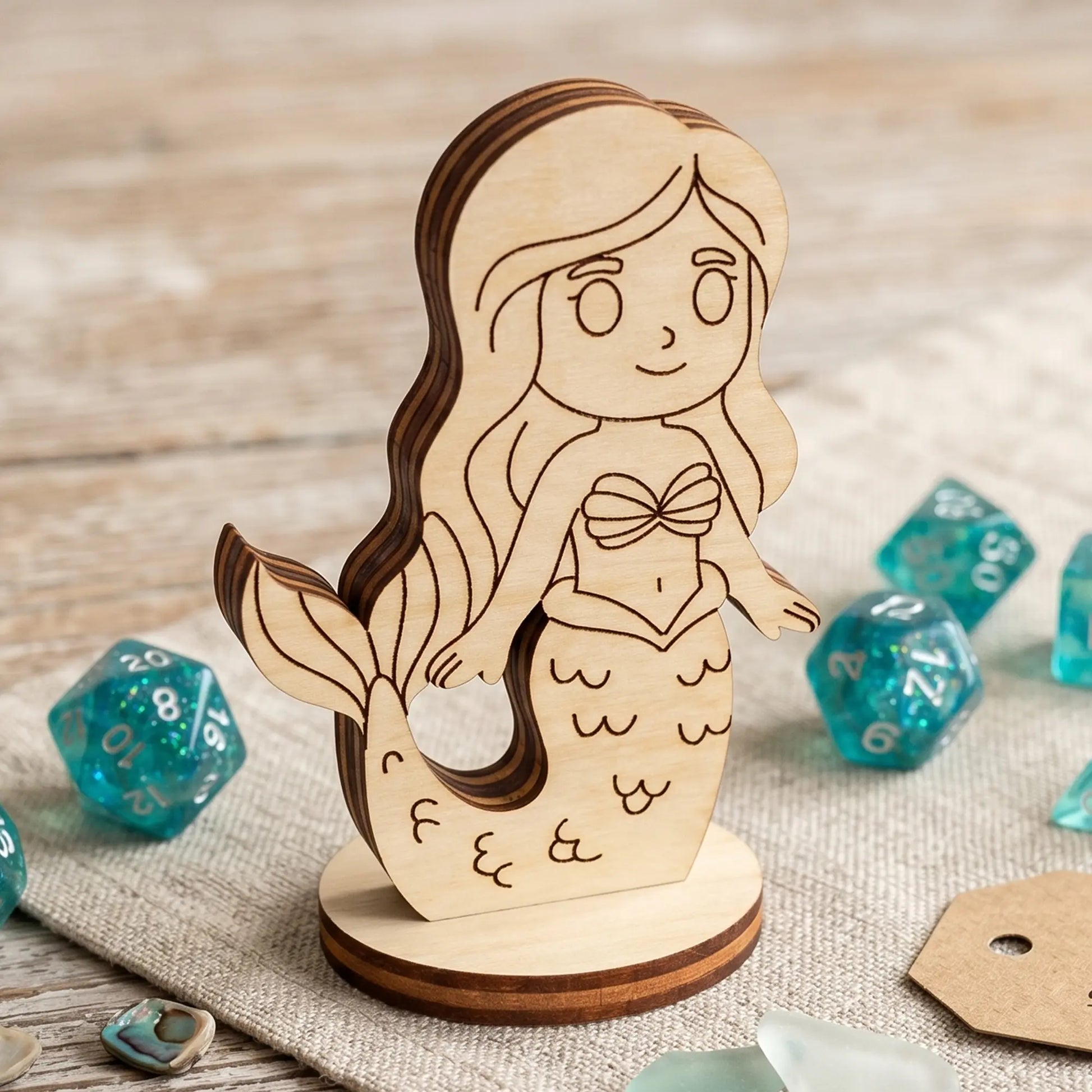 Detailed close-up of a laser cut plywood mermaid figurine with wood grain layers, standing on linen near teal glittery dice.